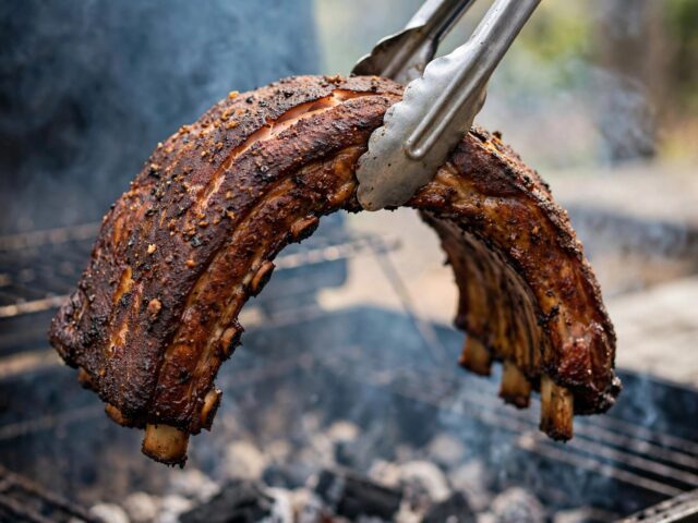 Rack of smoked pork ribs bending in a U-shape held by tongs over the grill