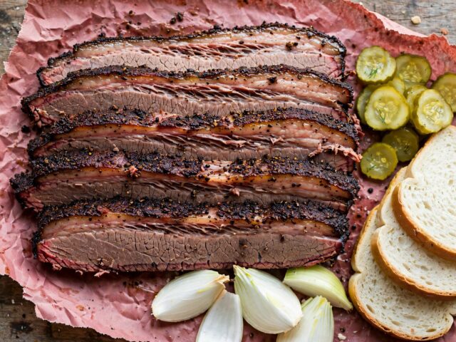 Sliced Central Texas brisket with dark peppery bark on pink butcher paper with pickles, white bread, and onion
