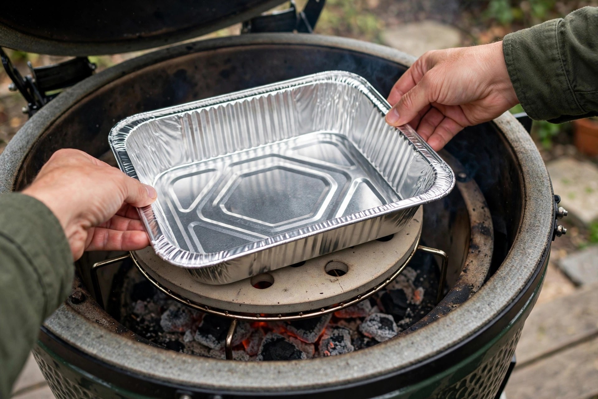 Hands placing an empty aluminum water pan on the heat deflector of a kamado grill before a cook